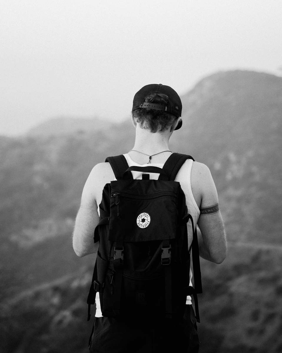 black-and-white man standing facing mountains wearing backpack backpack