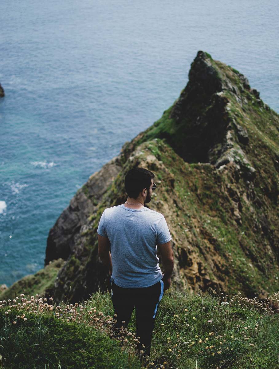 person man standing near cliff while facing body of water at daytime cliff