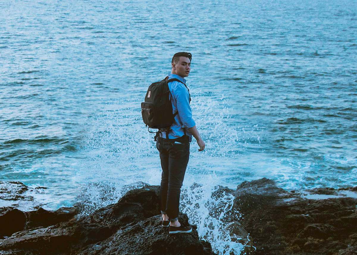 people man standing on gray rock formation near body of water wearing backpack person