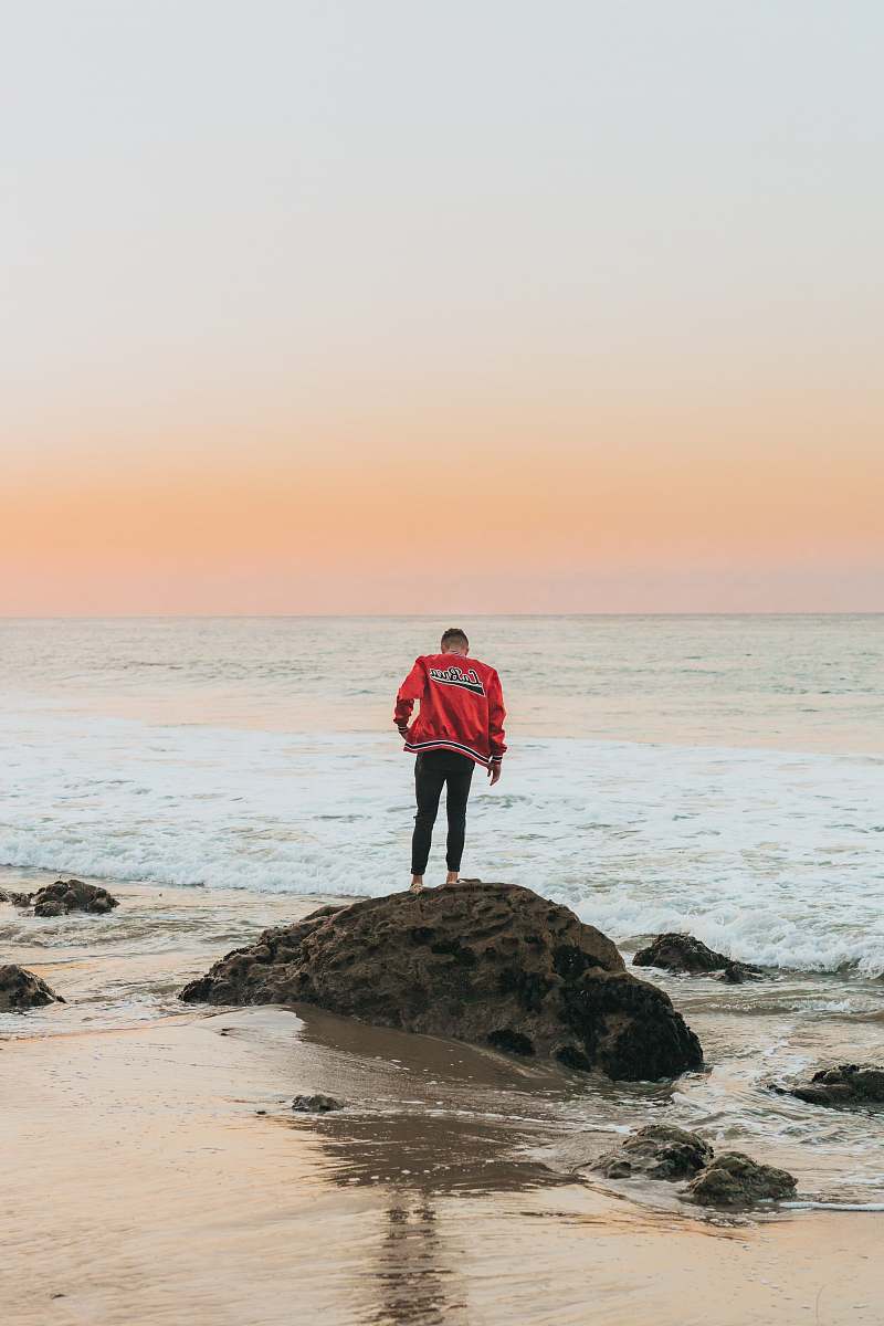 person man wearing orange jacket standing on seashore rock near beach people
