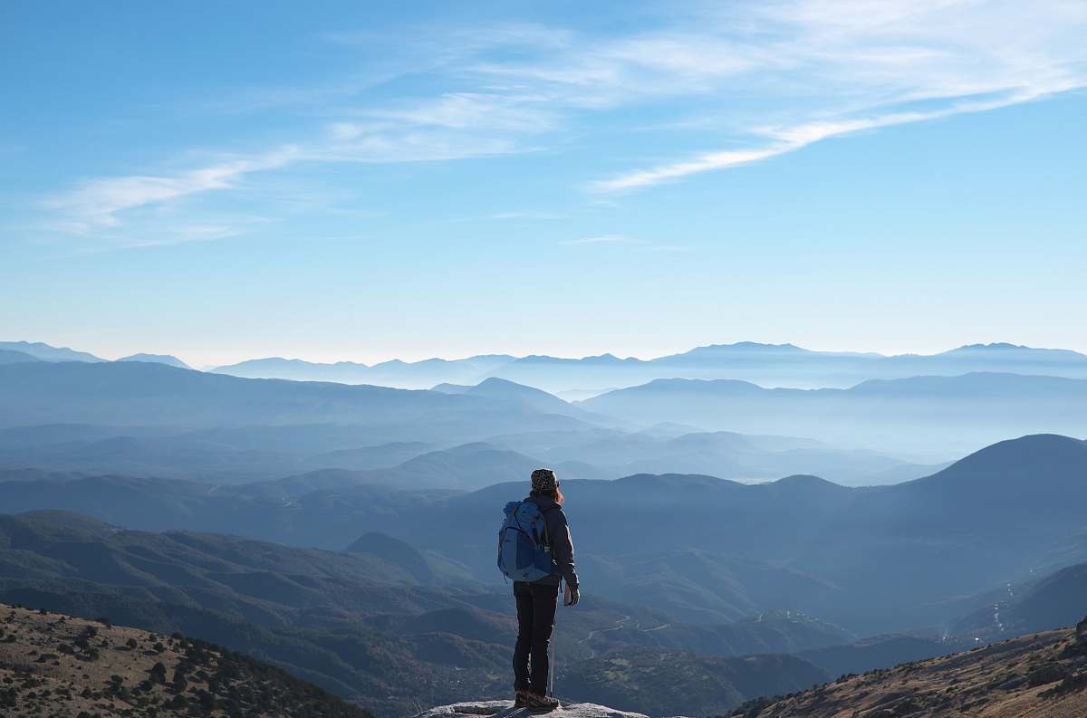 person person wearing backpack standing on cliff in front of mountains during daytime mountain range