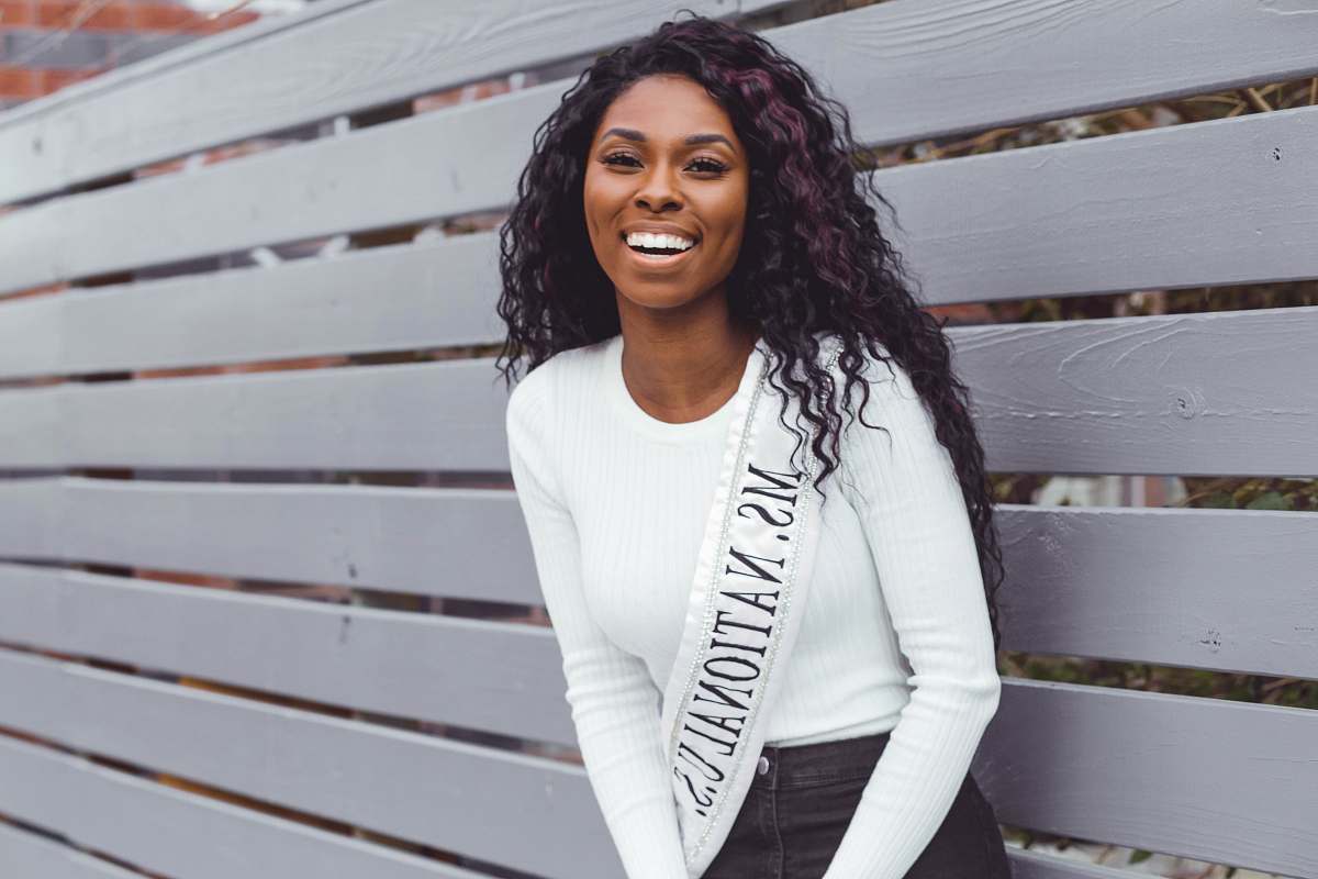 person smiling woman wearing Ms. National U.S. sash leaning on pallet wall people