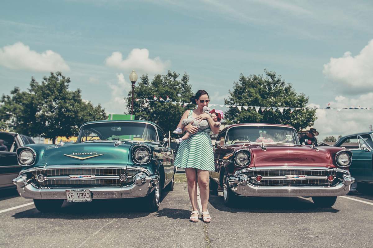 person woman carrying child standing between two vintage cars car
