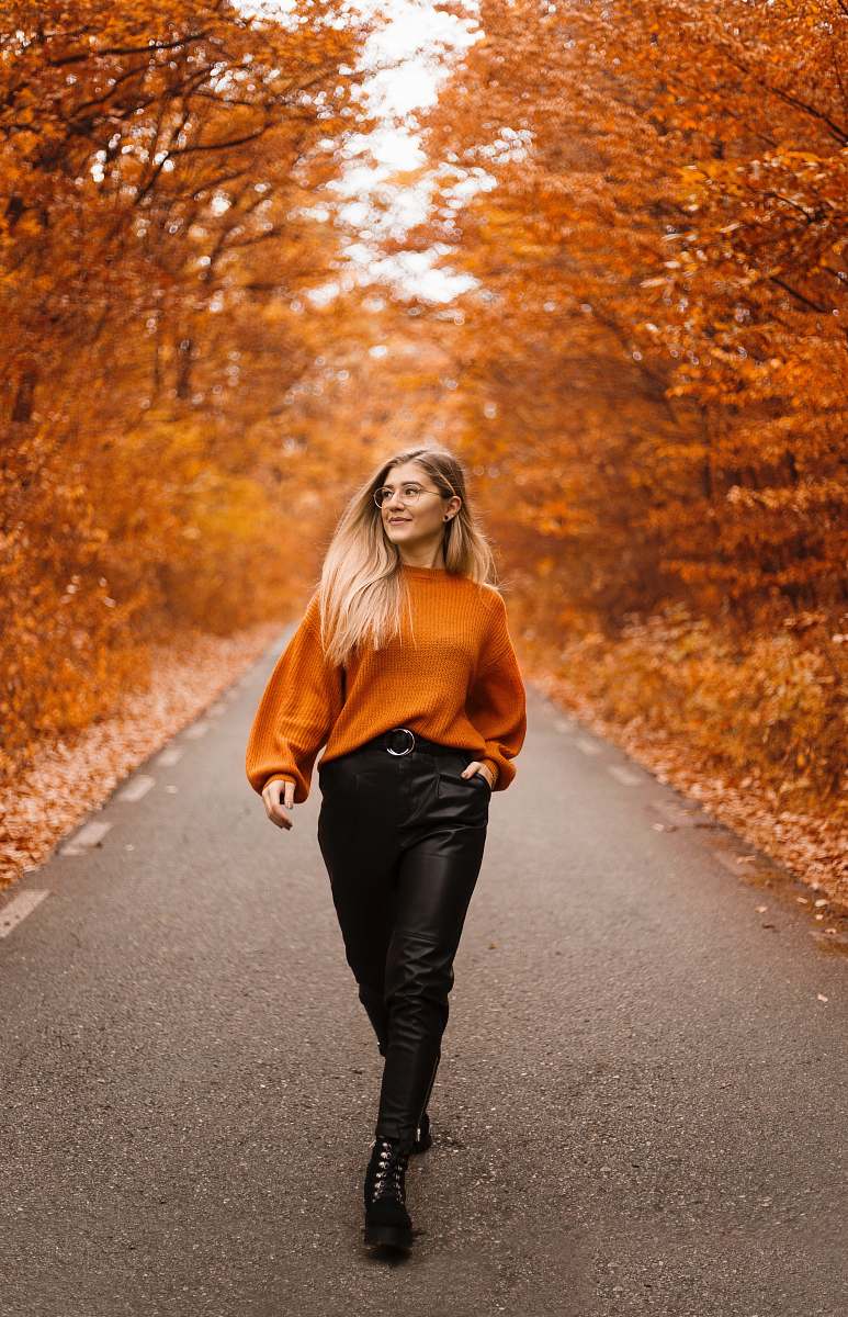 people woman in orange sweater and black pants walking on street between orange trees person