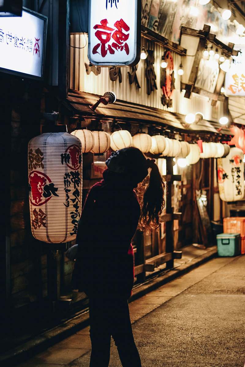 This captivating image captures a woman confidently walking down a city street at night, her gaze directed upwards towards the towering buildings. The scene is illuminated by soft streetlights, creating a vibrant urban atmosphere filled with possibilities. Ideal for projects related to city life, nightlife, fashion, and women's empowerment, this image embodies the essence of modern living and exploration.