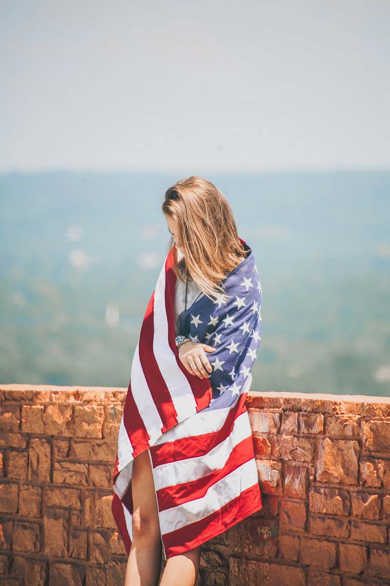 people woman standing leaning on brick wall wrapping body with U.S. flag near body of water person