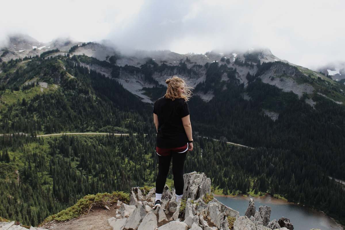 mountain woman standing on rock cliff overlooking trees and body of water with mountains at distance during daytime outdoors