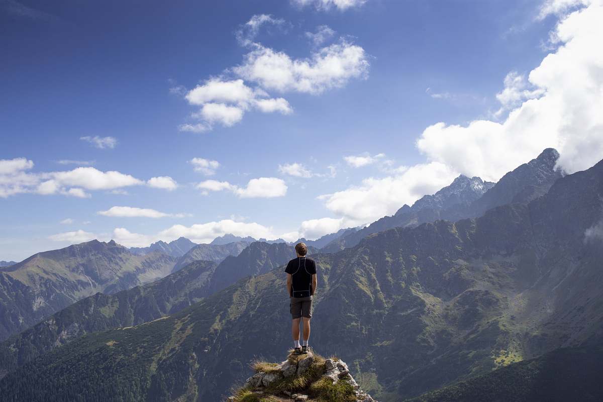 mountain man wearing black shirt and gray shorts on mountain hill beside mountains under white and blue cloudy skies mountain range