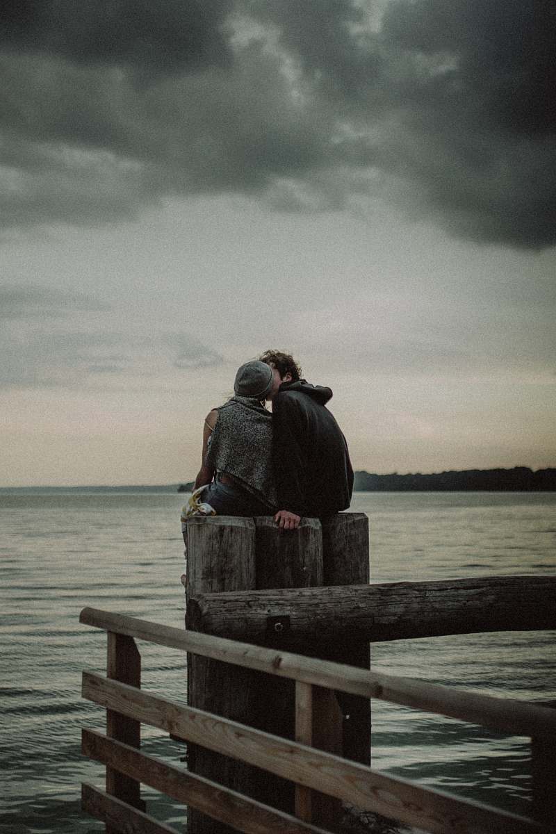 human man and woman sitting and kissing on post near the body of water person