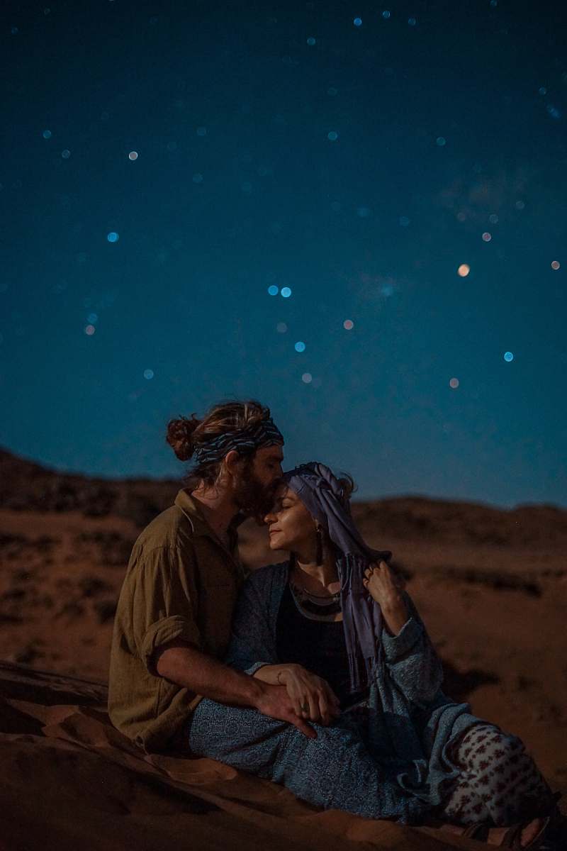 person man and woman sitting on desert sand under blue sky during nighttime human