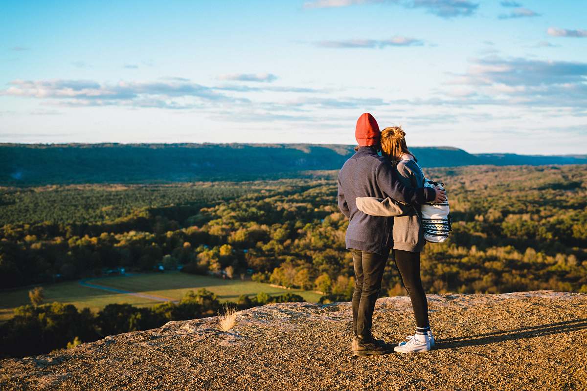human man and woman standing on mountain cliff in front of green trees person