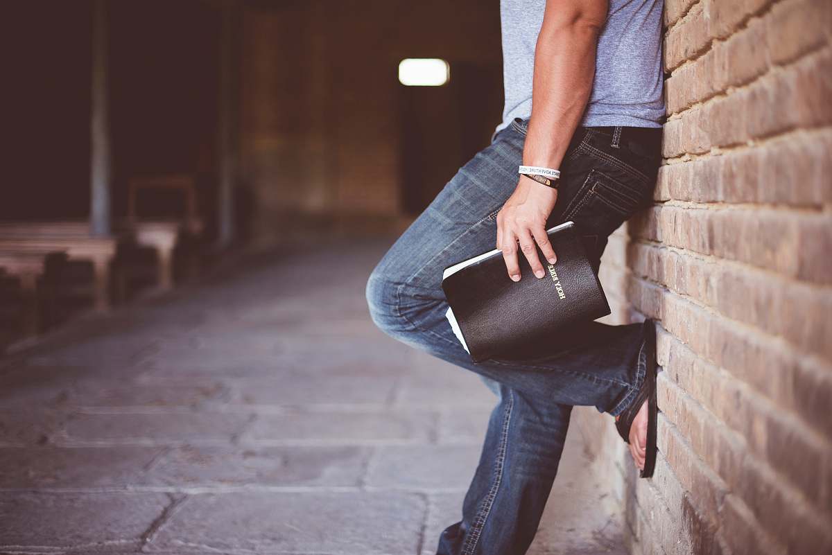 person man holding Holy Bible leaning on bricked wall book
