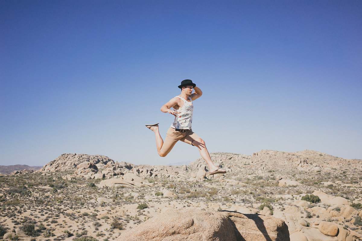 person man jumping on brown stone while holding his hat desert