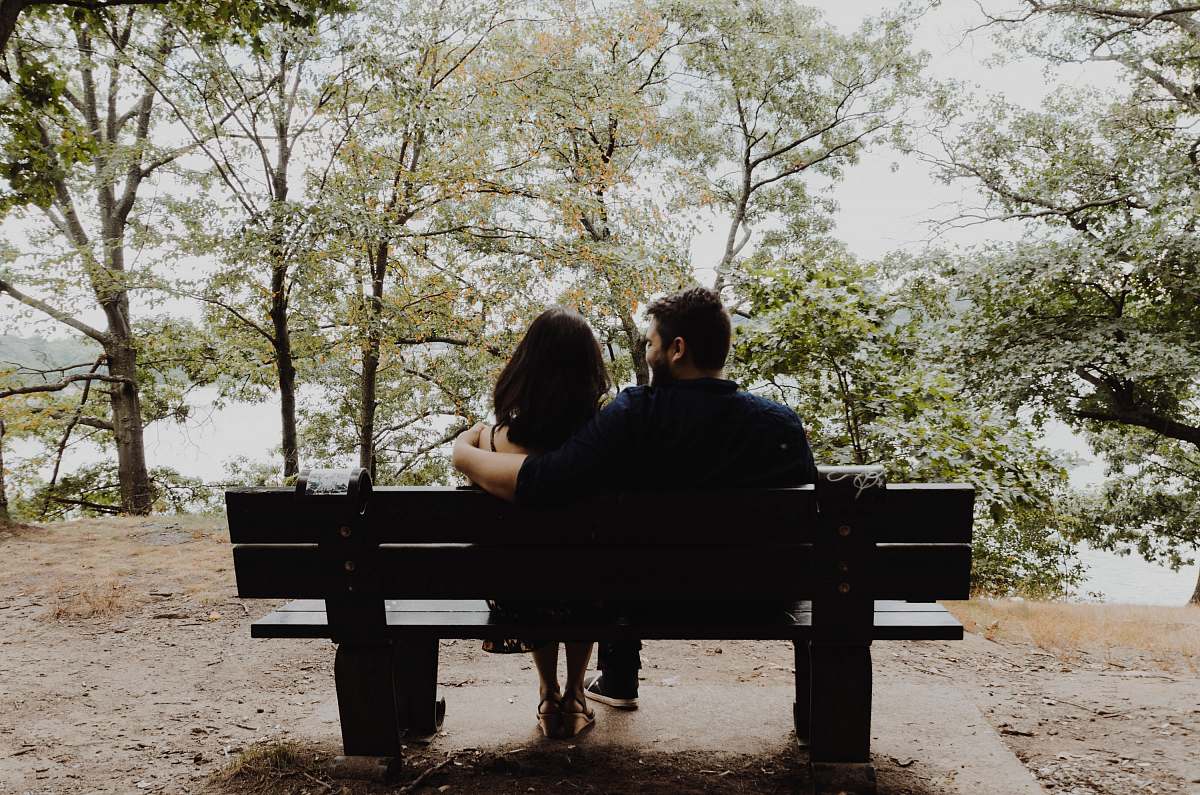 human man looking to woman sitting on black wooden bench in front of tall trees during daytime bench