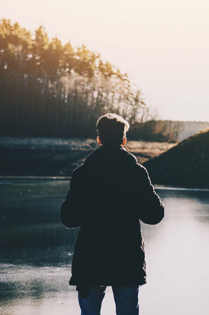 human man standing infront of lake and trees during daytime person