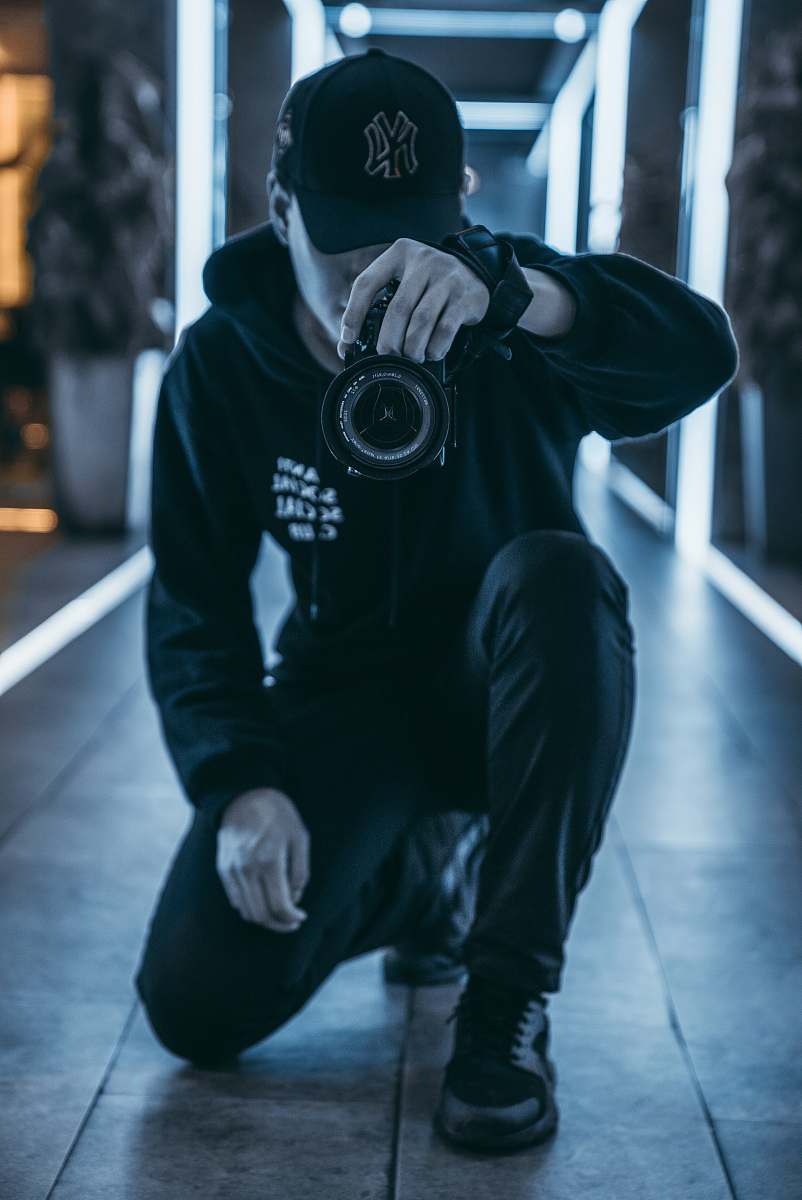 A focused man kneels on the floor of a shadowy room, holding a camera poised to capture the perfect shot. The ambiance is filled with creative energy, making this image ideal for photography enthusiasts, marketing materials, or any project that celebrates the art of capturing moments. The interplay of light and dark in the scene adds a dramatic touch, evoking curiosity and intrigue in viewers.