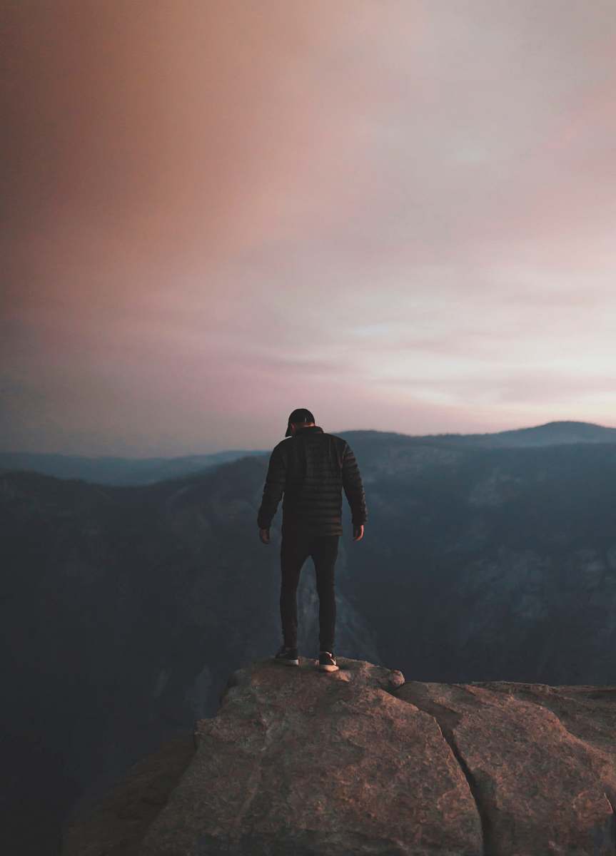 human man wearing black bubble jacket on rocky mountain hill in front of mountains under white, pink, and blue cloudy skies during daytime person
