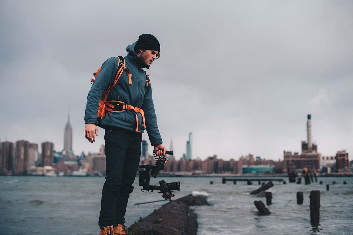 human man wearing teal zip-up bubble jacket taking photo of body of water with camera with stabilizer person