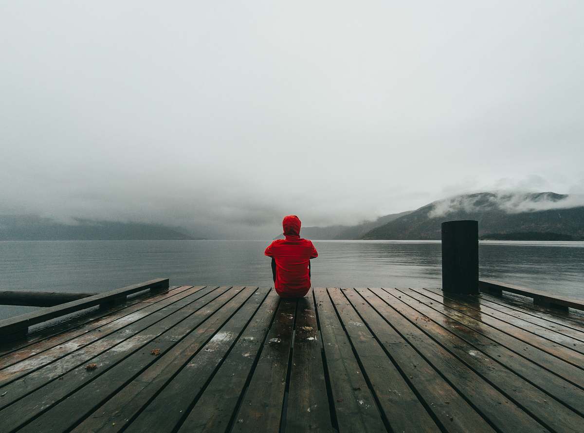 human person wearing red hooded jacket sitting on brown and black wooden sea dock near mountains under gray sky during daytime person
