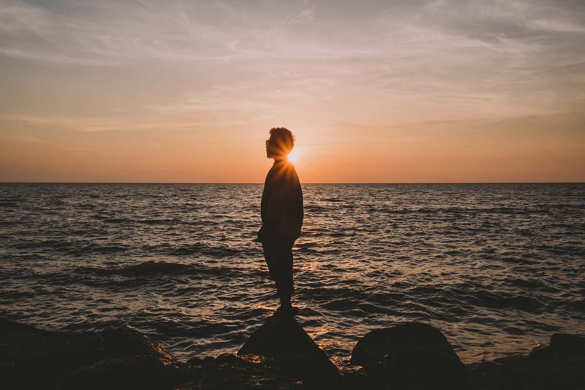 human silhouette of man standing on stone beside wavy beach water during sunset person