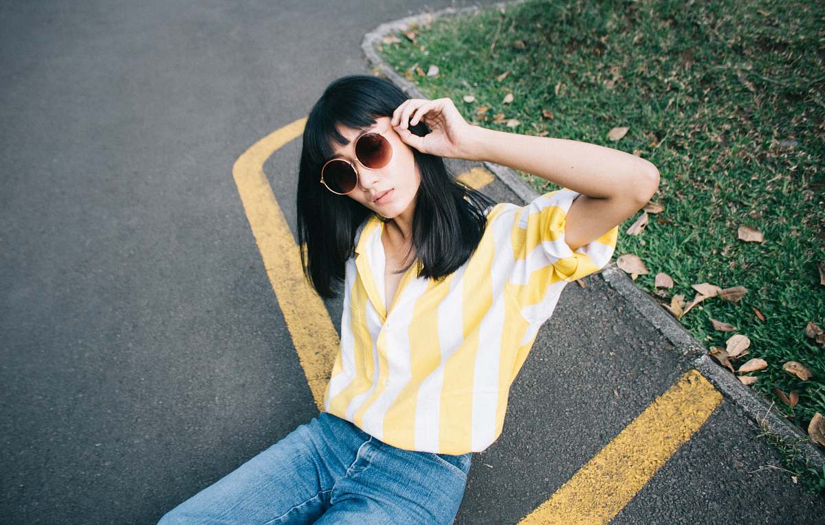 person woman wearing yellow and white striped collared shirt holding round-shaped brown sunglasses sitting on gray concrete pavement during daytime human