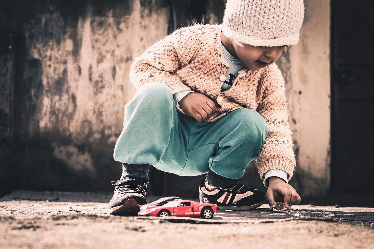 people child squatting on beige floor holding rock near red sports coupe scale model at daytime human