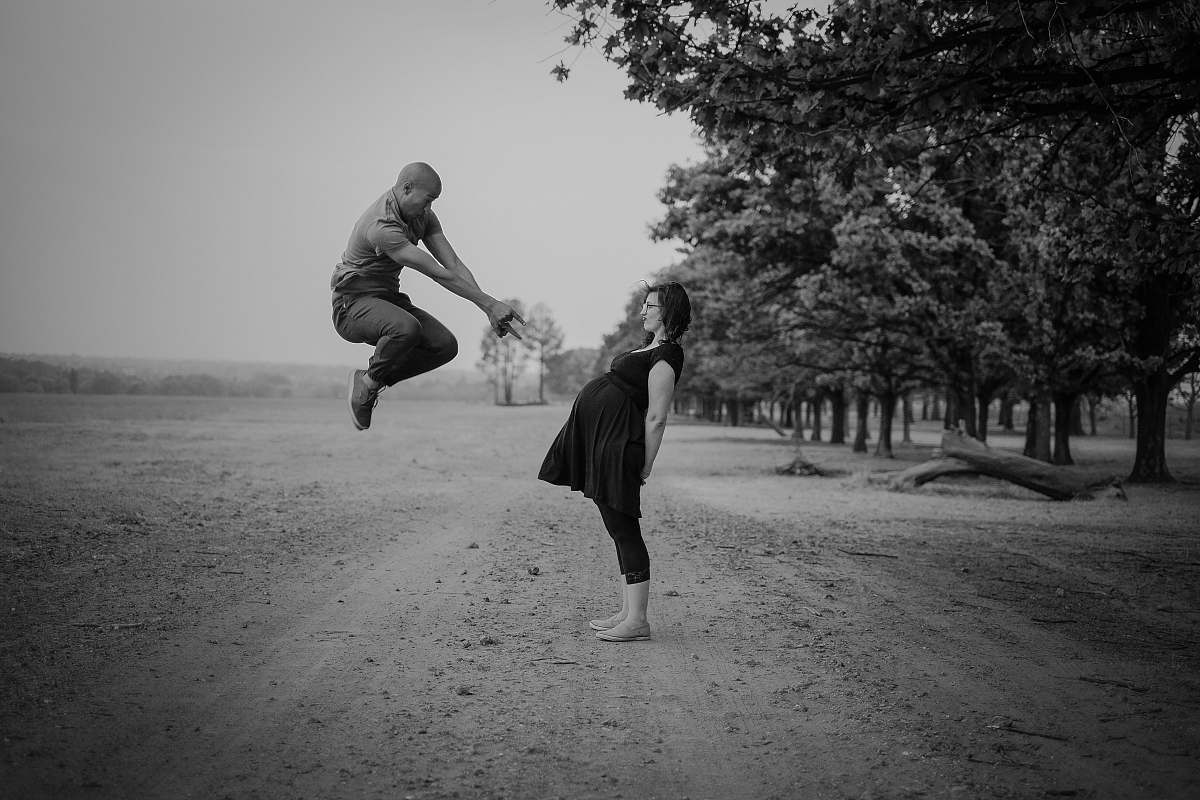 human grayscale photo of man jumping in front of pregnant woman near trees during daytime black-and-white