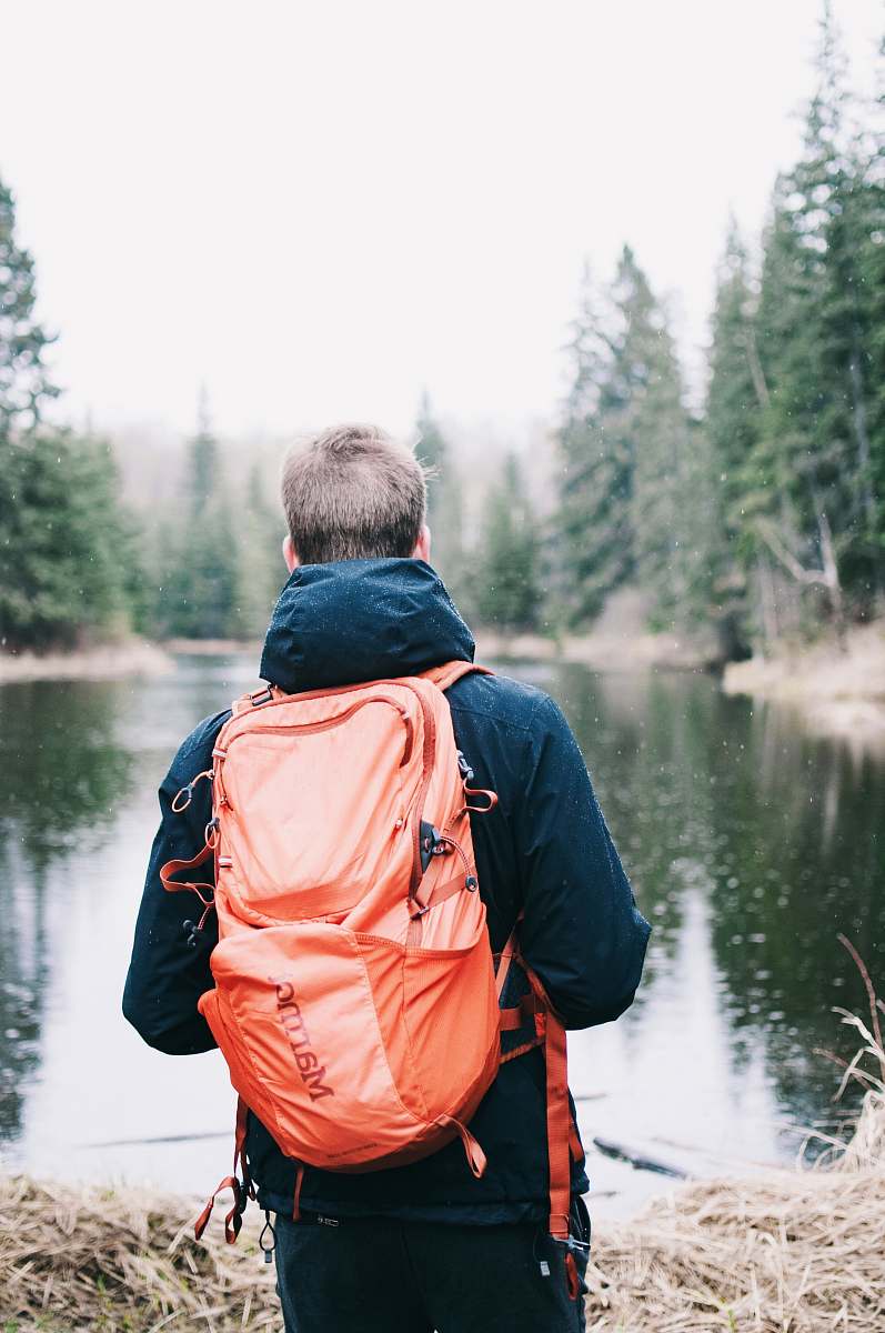 human man wearing blue jacket and red backpack facing body of water back