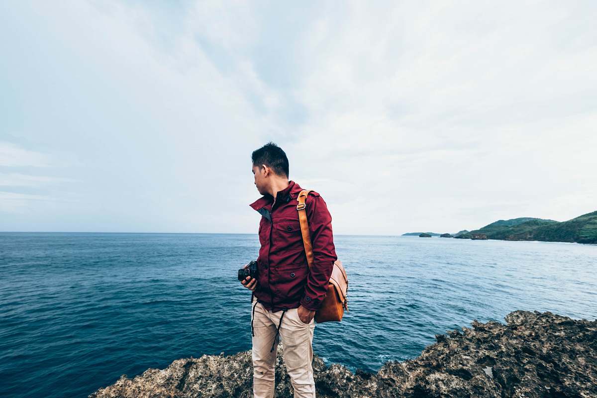 human man wearing red zip-up jacket standing on top of rocky cliff beside body of water during daytime people
