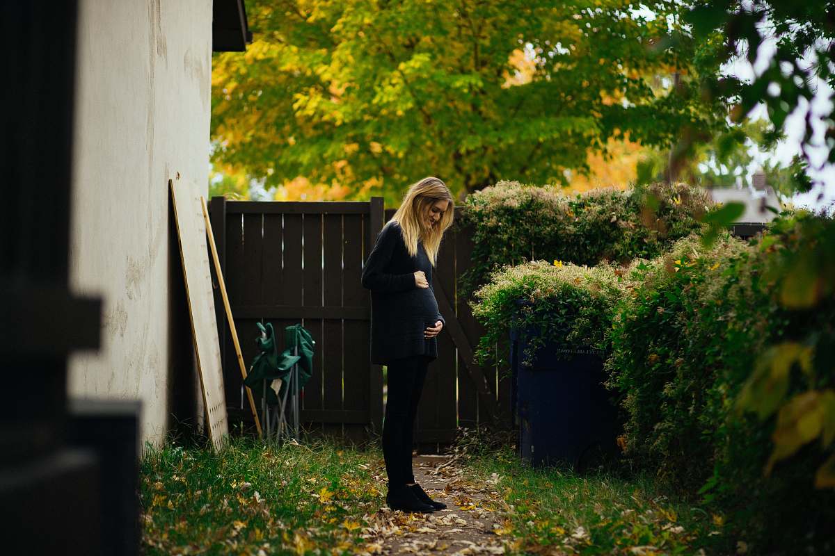 human pregnant woman touching belly while looking at green bush near brown wooden fence during daytime people
