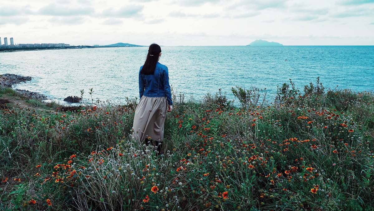people woman in blue blouse and gray skirt standing on red flower field beside body of water during daytime human