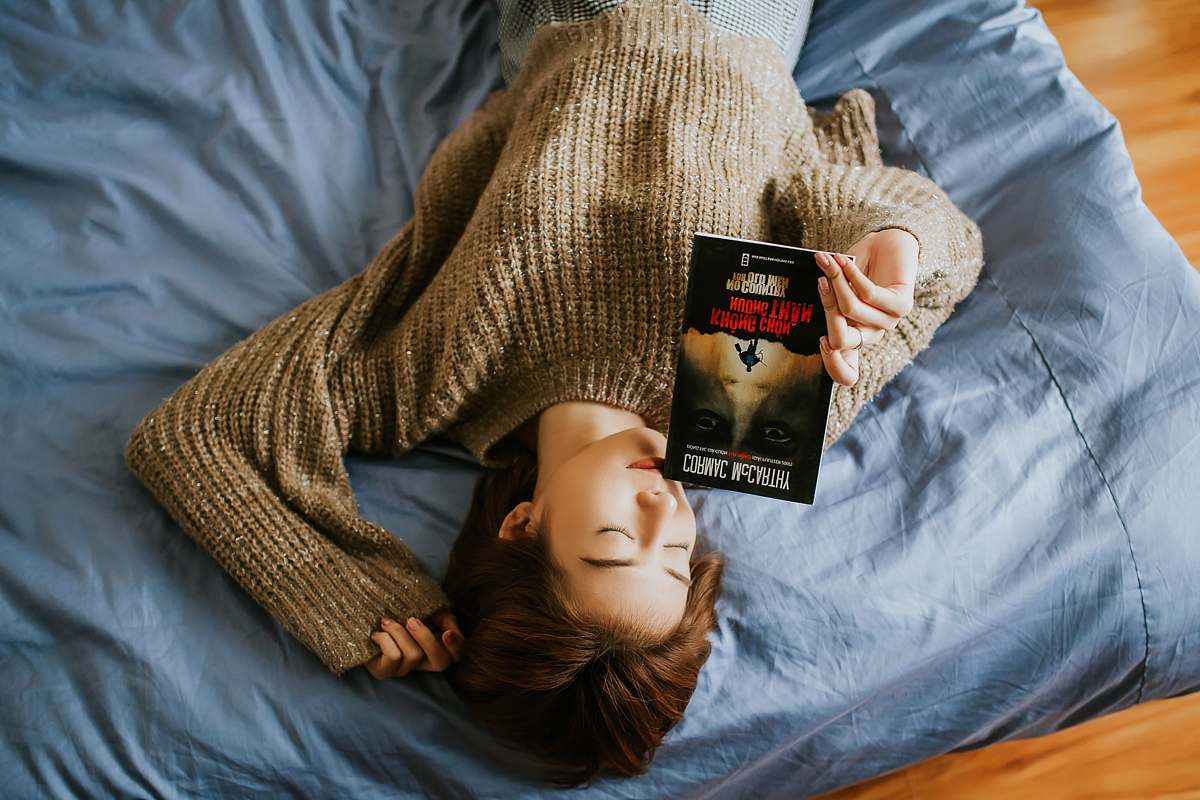 people woman in sweater holding book while lying on bed human