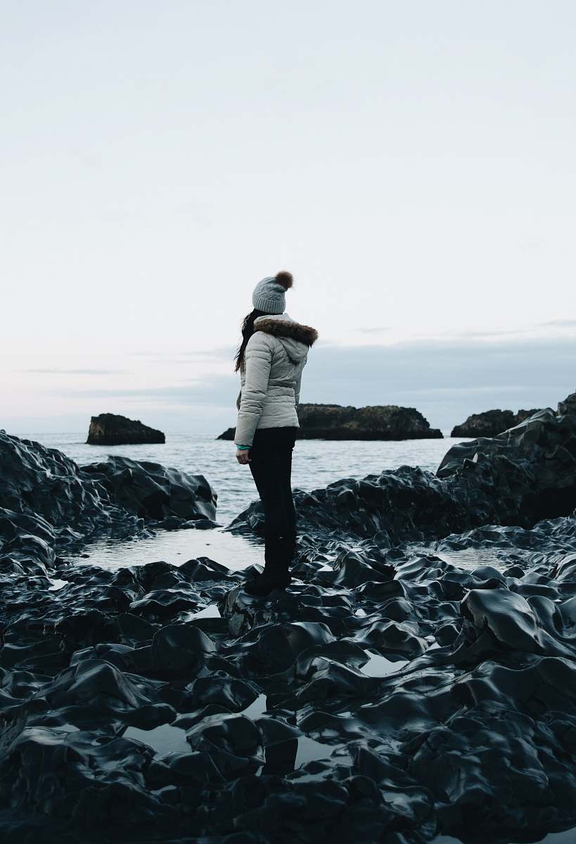 people woman in white parka jacket and black pants outfit standing on rocks near body of water under white sky at daytime human