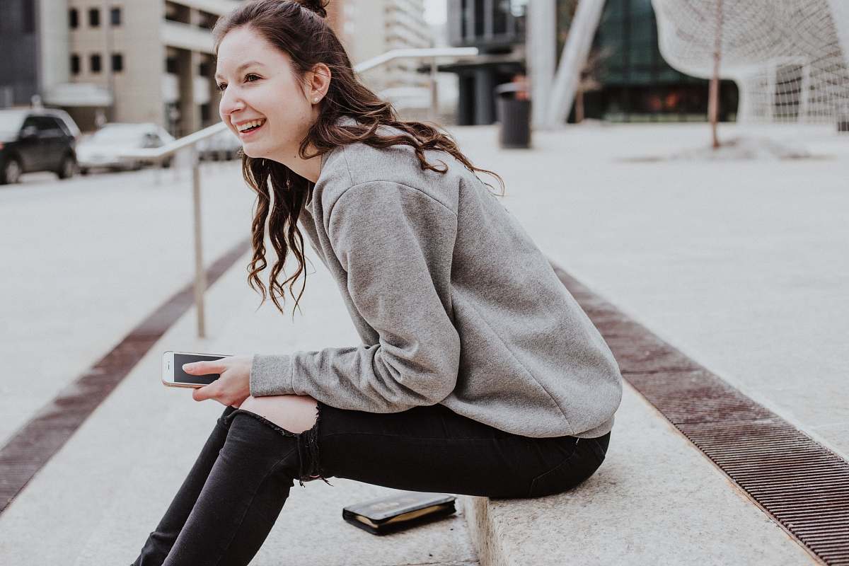 people woman sits on sidewalk holding smartphone while smiling human
