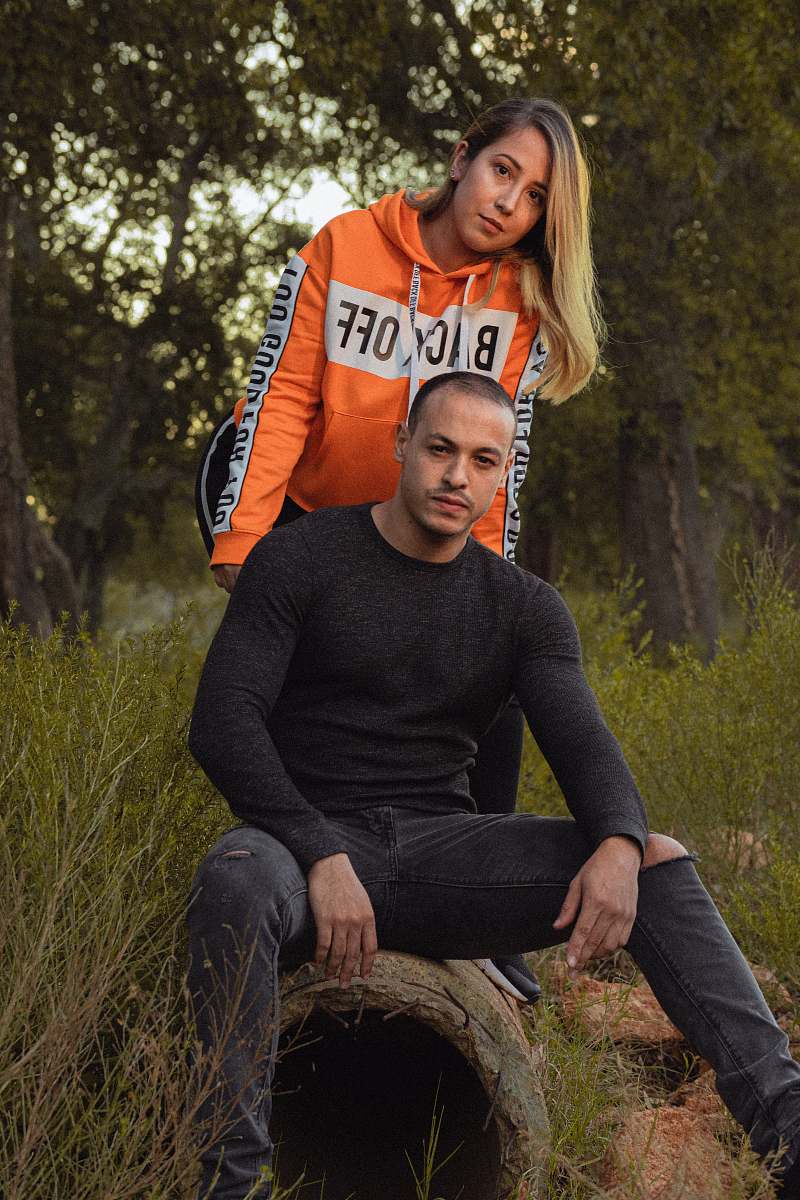 apparel man sitting on bench beside woman standing surrounded with trees sweater