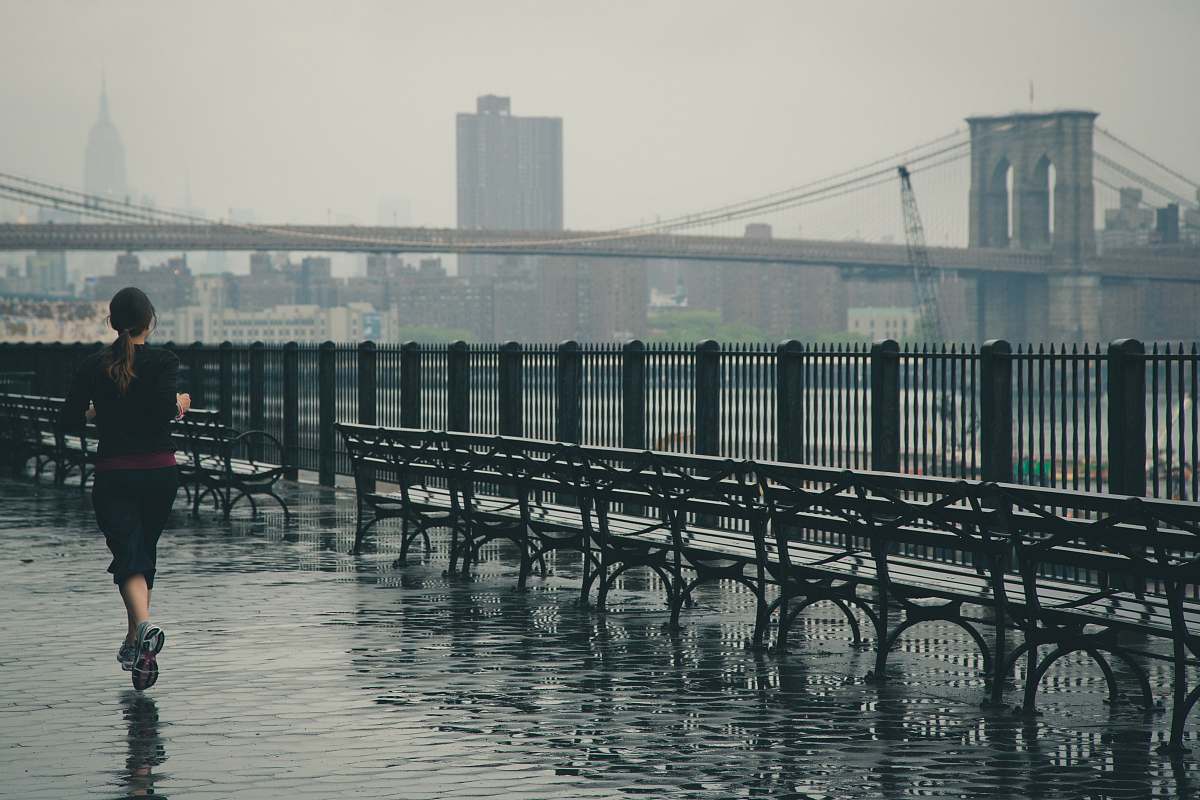 Experience the energy of an athletic woman running alongside a serene body of water, with the iconic Brooklyn Bridge majestically looming in the background. This vibrant scene captures the essence of urban fitness and resilience, illustrating a perfect blend of nature and city life. Ideal for lifestyle blogs, fitness articles, and motivational content, this image showcases the beauty of exercising outdoors, even in rainy conditions.