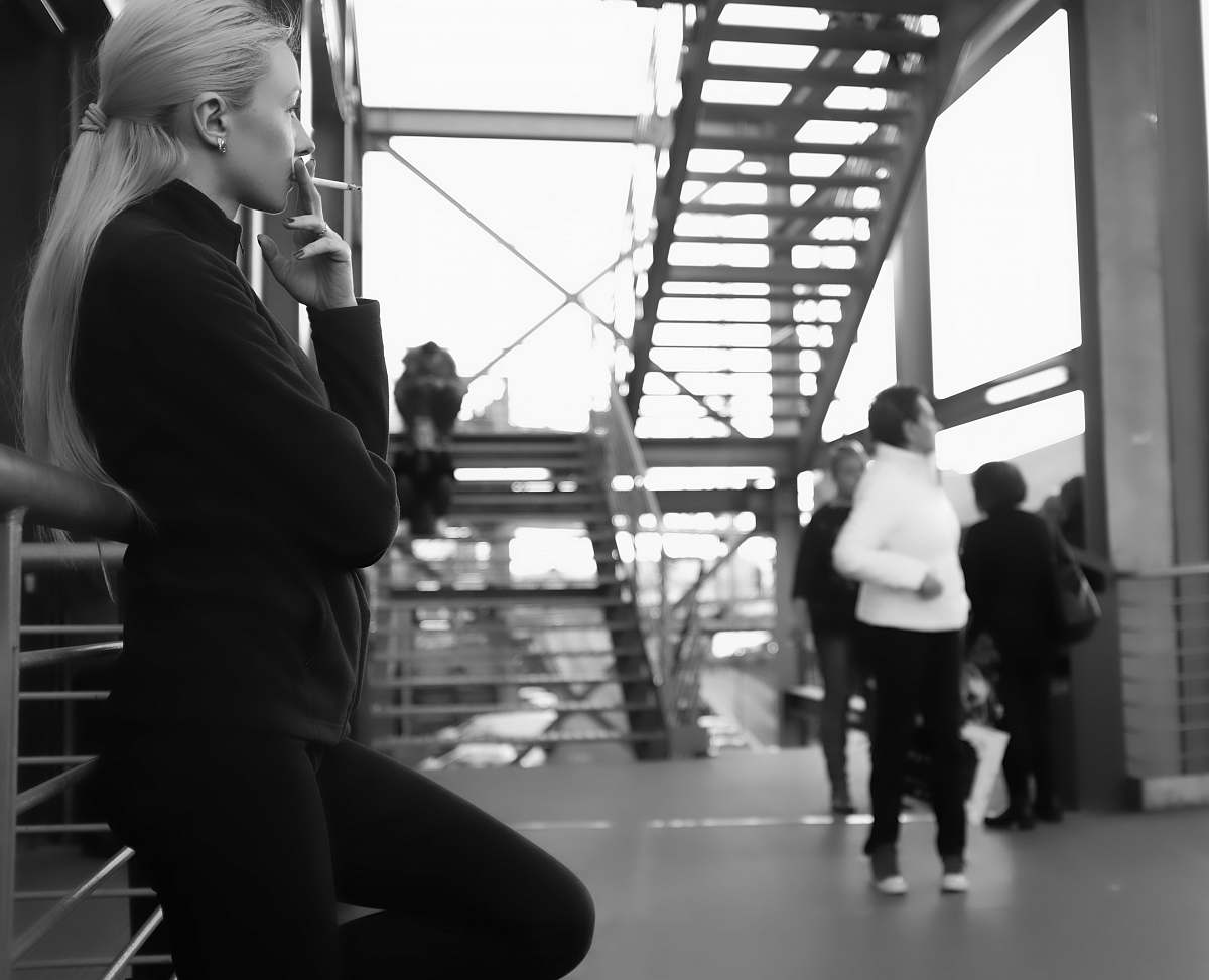 person grayscale photography of woman using cigarette while leaning on metal rail black-and-white