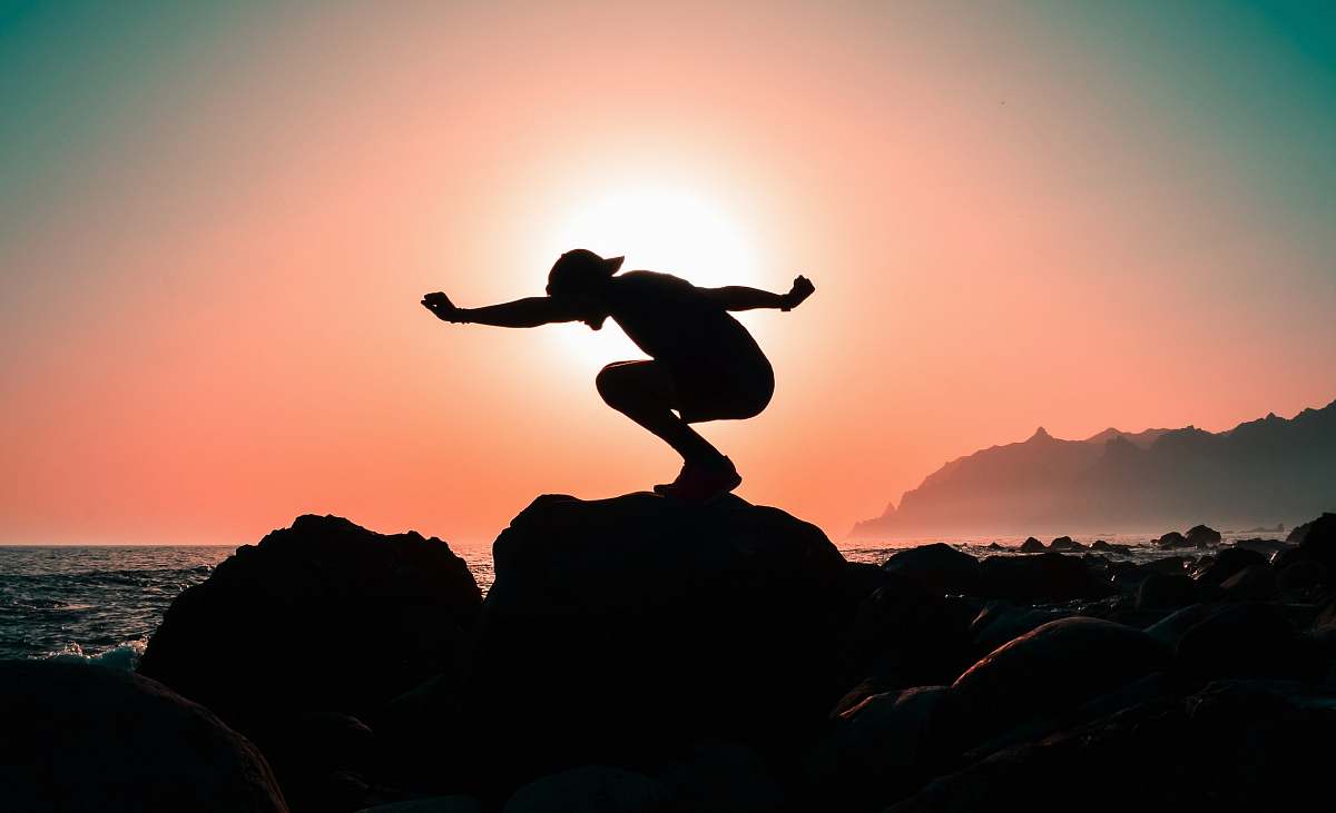 person silhouette photography of person squatting on rock near body of water during golden hour nature