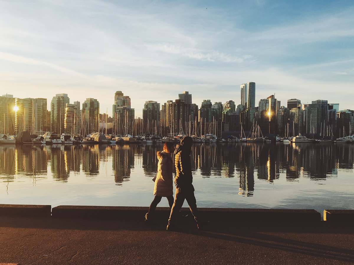 person two people walking near body of water viewing city with high-rise buildings under white and blue sky during daytime building
