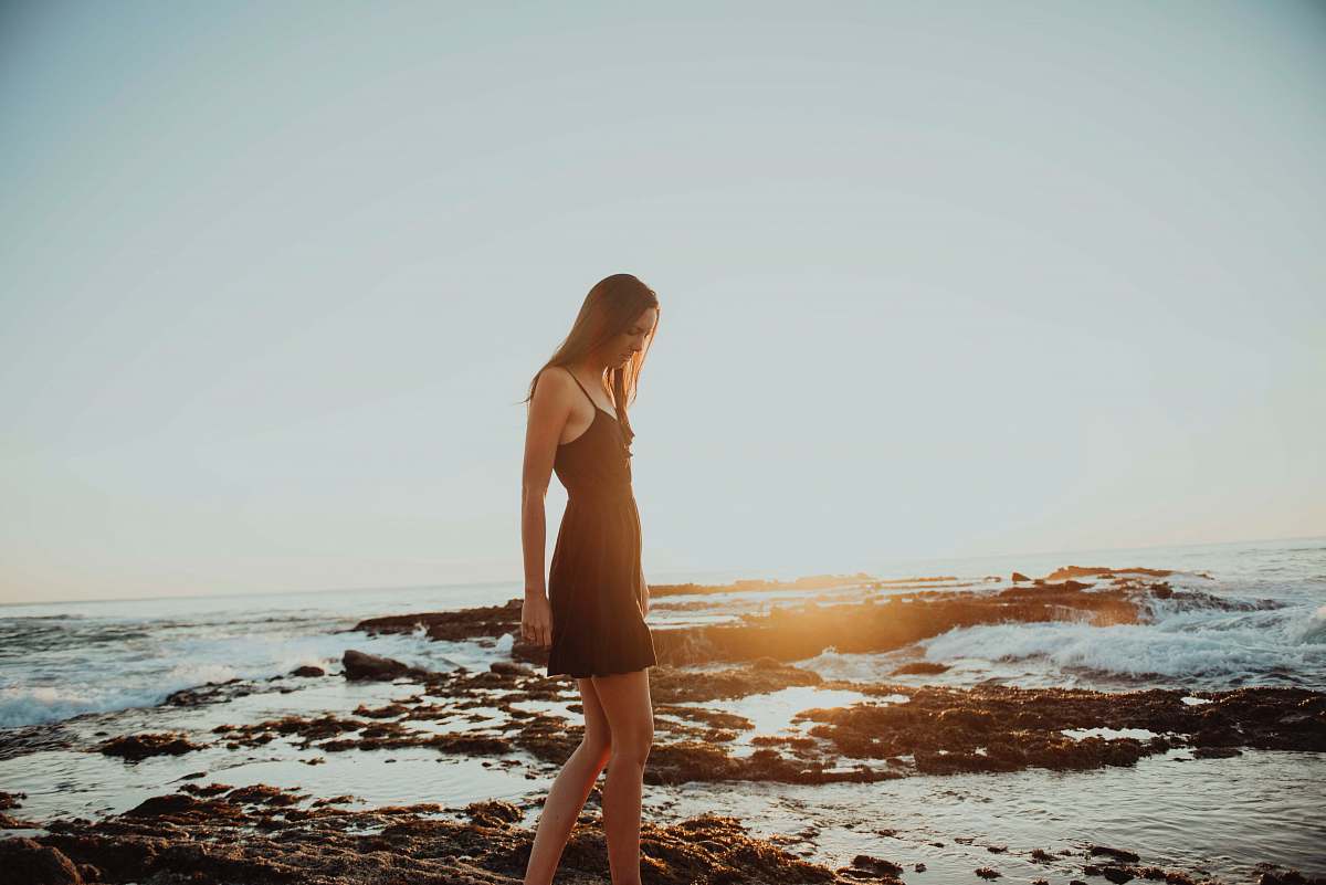 people woman in black spaghetti strap dress walking on rock formation surrounded by body of water during daytime person