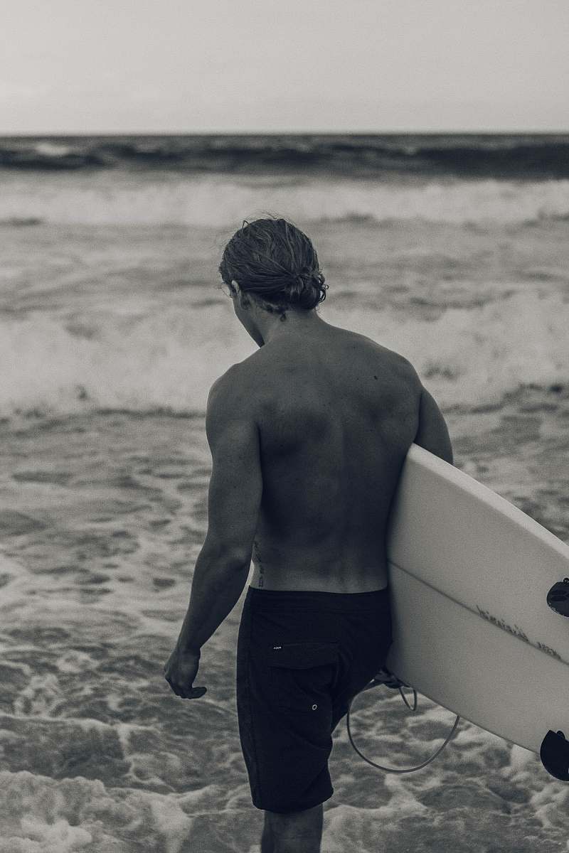 outdoors grayscale photography of man carrying a surfboard by the beach sea