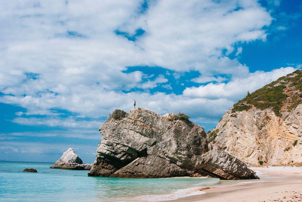 beach man standing on top on rock formation surrounded by body of water during daytime coast