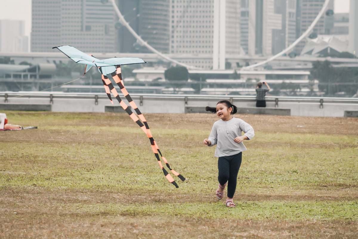 human girl swearing gray crew-neck long-sleeved shirt running in green field near orange and white plane kite viewing high-rise buildings sport