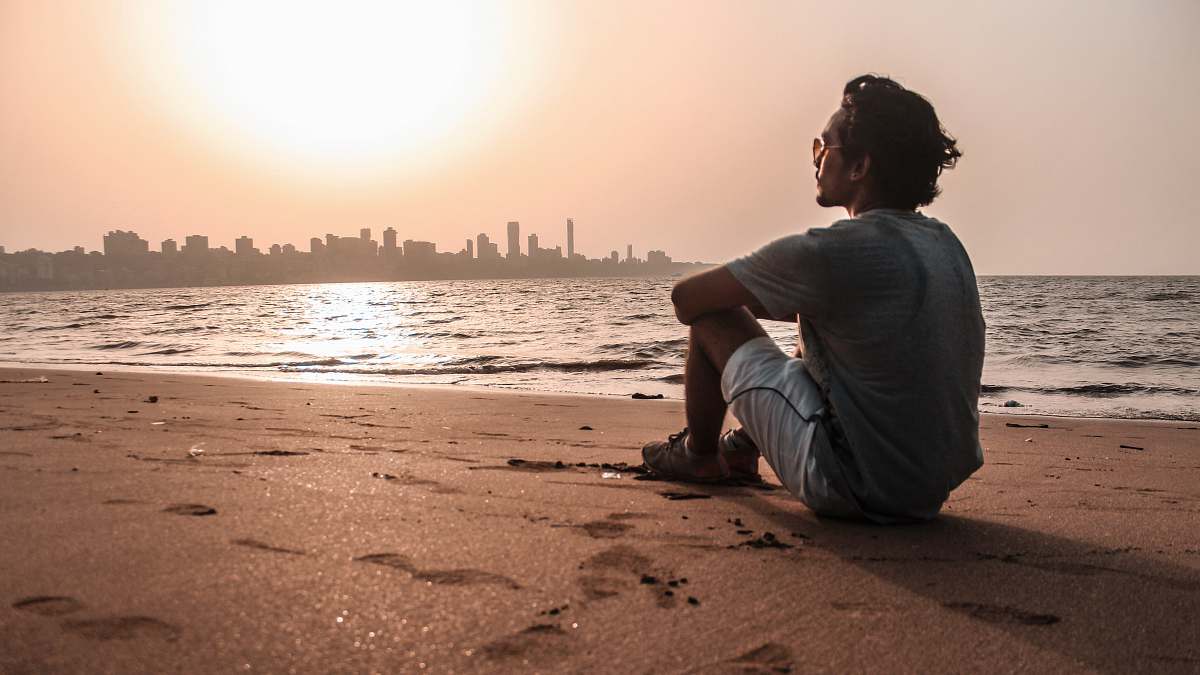 human man in gray shirt sitting on sand near body of water sitting