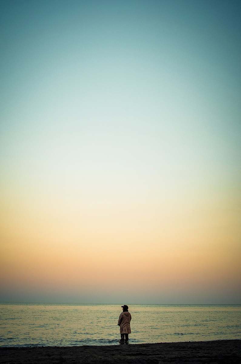 A solitary man stands near the tranquil shore, absorbing the breathtaking views of a vibrant sunset. The sky is painted with hues of orange, pink, and purple, creating a picturesque backdrop that embodies peace and reflection. This evocative scene captures the essence of solitude and nature, making it ideal for projects related to personal growth, relaxation, travel, and outdoor lifestyle.