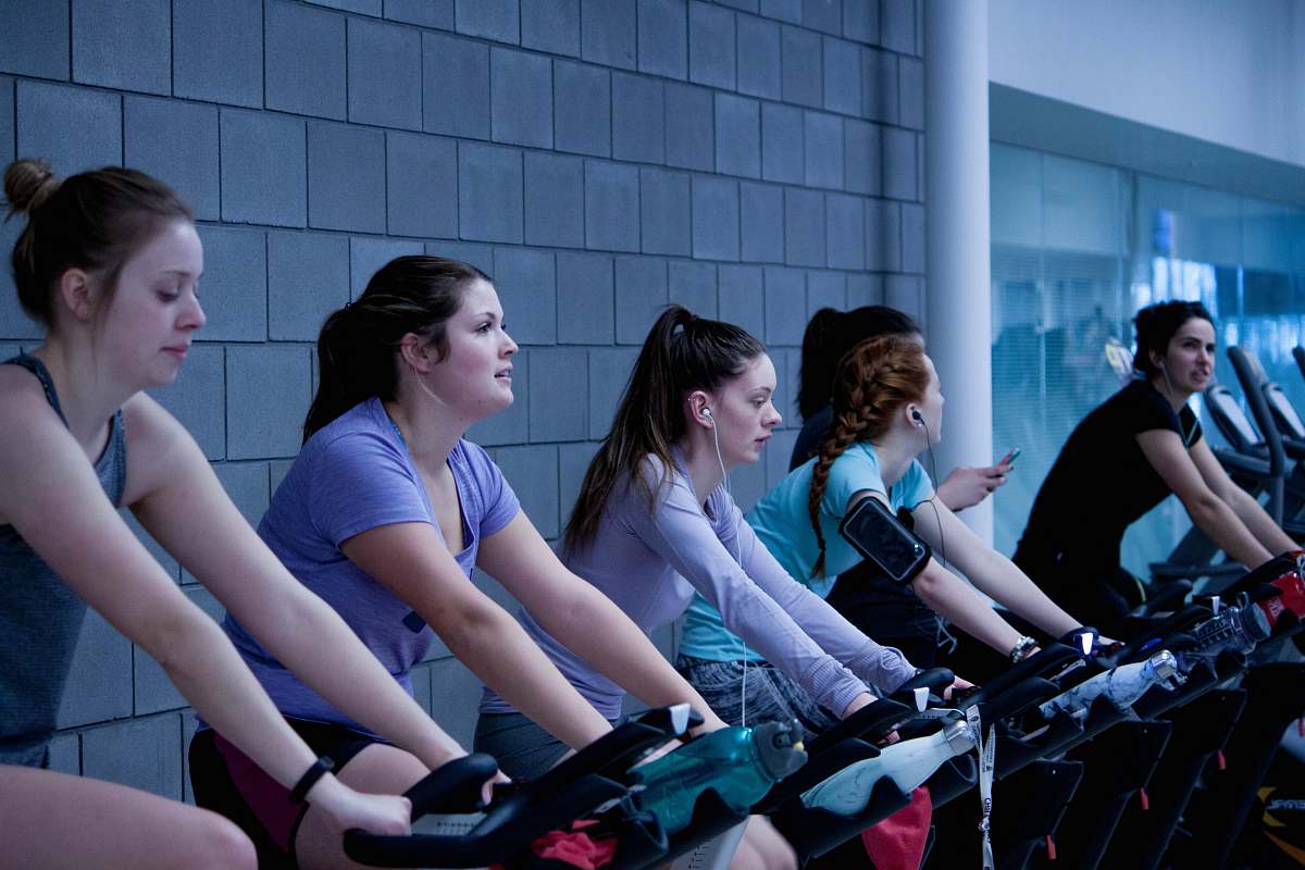 human women taking exercise on black stationary bikes in front of gray concrete wall people