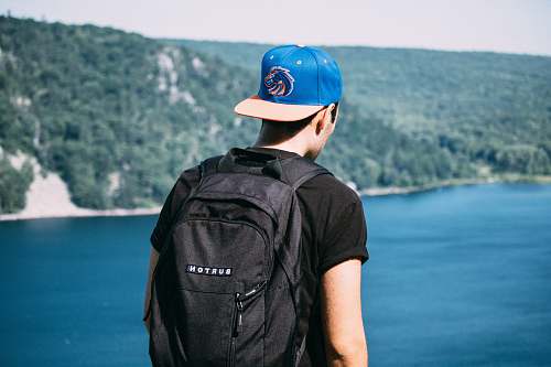 person man wearing black t-shirt, black backpack, and blue fitted cap facing body of water photo human