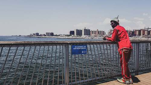 human man in red shirt standing and leaning on metal railings near sea during daytime person
