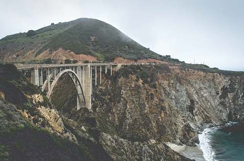 bridge grey concrete bridge between brown and black cliff near body of water during daytime arch