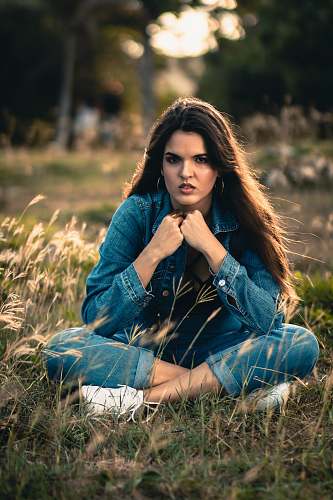 apparel woman in blue denim jacket and blue denim jeans sitting on green grass field during daytime female